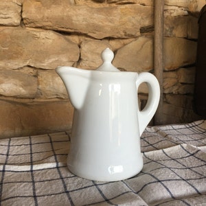May include: A white ceramic pitcher with a lid and a curved handle. The pitcher is on a checkered cloth, with a stone wall in the background. The pitcher has a spout for pouring.