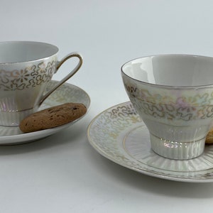 May include: Two white teacups and saucers with gold and white floral patterns. Each saucer has a cookie on it.