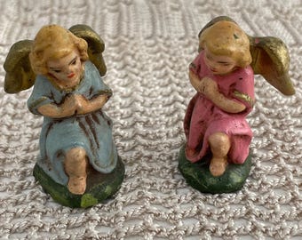 Op de afbeelding: Twee kleine engelfiguurtjes in een knielende, biddende houding. De ene engel draagt een lichtblauw gewaad en de andere een roze gewaad. Beide figuren hebben blond haar, goudkleurige vleugels en staan op kleine, donkergroene, ronde voetstukken.