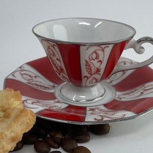 May include: A red and white porcelain teacup and saucer set with a floral design. The saucer has a scalloped edge and the cup has a silver handle.