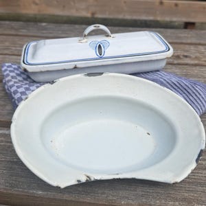 May include: A white enamel bedpan and a matching lidded container with blue accents. The bedpan is oval-shaped with a curved edge. The container has a handle and a blue floral design. Both items rest on a blue and white striped cloth.