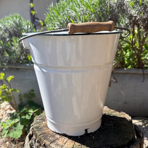May include: A white enamel bucket with a dark rim and a wooden handle. The bucket has a metal wire handle and is sitting on a tree stump. The bucket is a classic design, suitable for various uses.