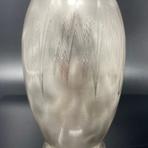 May include: A silver vase with an oval body and a textured surface. The surface has a repeating zigzag pattern. The base is circular and wider than the body. The top is smooth and open. The vase is approximately 20 cm tall.