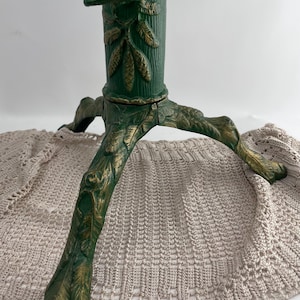 May include: A green and gold metal Christmas tree stand featuring a cylindrical center column with a single tightening screw and three legs. The surface is decorated with embossed pine cone and needle patterns throughout the entire structure.
