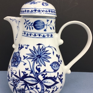 Op de afbeelding: Een witte porseleinen koffiekan met een blauw en wit bloemenpatroon. De kan heeft een deksel en een handvat.