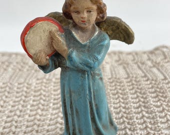 Op de afbeelding: Een kleine, vintage figuur van een engel in een vloeiend blauw gewaad, met een tamboerijn. De engel heeft bruin haar en goudkleurige vleugels. De tamboerijn heeft een rode rand en een crèmekleurig midden. Het beeldje staat op een crèmekleurig, getextureerd oppervlak.