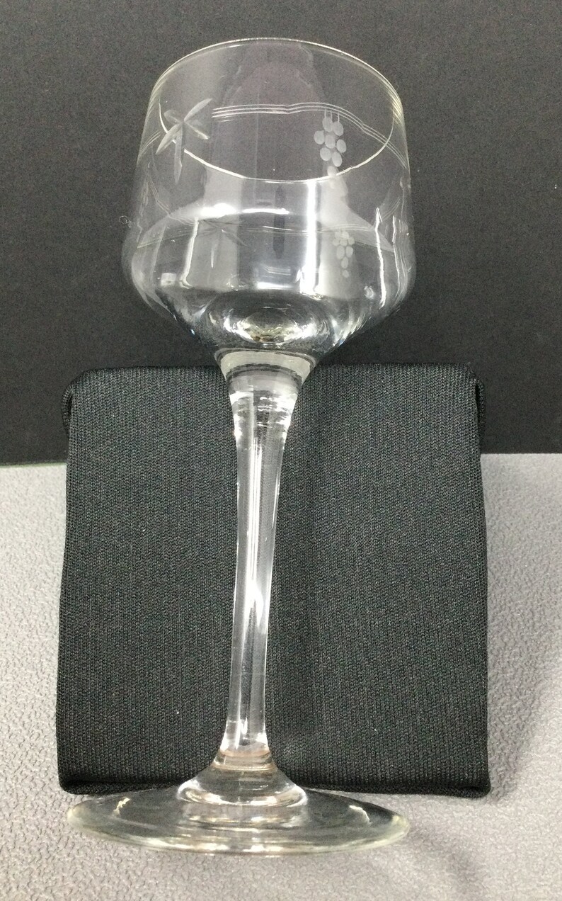 May include: A clear glass wine glass with a long stem and a wide bowl. The glass has an etched design of grapes and vines around the top of the bowl.