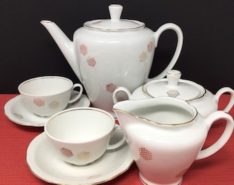May include: A white porcelain tea set with a gold trim and a geometric pattern in shades of red and gold. The set includes a teapot, creamer, sugar bowl, and two teacups with saucers.