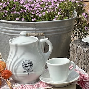 May include: White porcelain teapot with a lid and a handle. The teapot has a black and white label that reads "Bültner D.R. Patent". The teapot is sitting on a red and white checkered tablecloth.