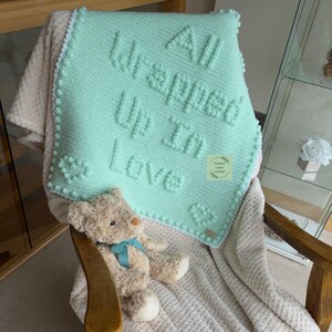 May include: A mint green crocheted blanket featuring the raised text "All Wrapped Up In Love" and two heart shapes, draped over a beige textured throw blanket and a teddy bear with a blue ribbon, all resting on a wooden rocking chair.