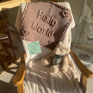Puede incluir: Una manta de bebé hecha a mano, tejida a ganchillo, de color rosa suave, con las palabras "Hello World" cosidas en la parte delantera. Un pequeño oso de peluche con una cinta verde está sentado sobre la manta, que está sobre una manta crema en una mecedora de madera.