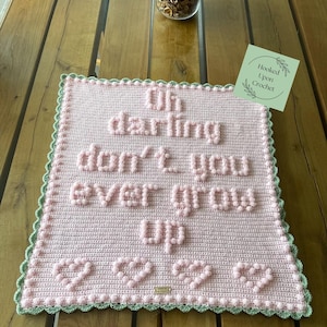 May include: A rectangular light pink crochet blanket featuring the raised bobble stitch text "Oh darling don't you ever grow up" and four hearts at the bottom. The blanket is finished with a scalloped sage green border and includes a small brand tag.