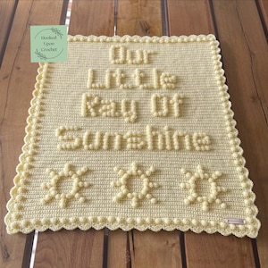 Puede incluir: Una manta de ganchillo amarilla clara hecha a mano con las palabras "Our Little Ray Of Sunshine" en letras en relieve. La manta presenta tres diseños de sol y un borde festoneado. Una pequeña etiqueta en la esquina inferior derecha dice "Hooked Upon Crochet".