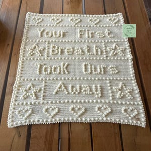 May include: A white crocheted baby blanket with the text "Your First Breath Took Our Aways" in raised letters. The blanket is decorated with small white pom-poms and stars.