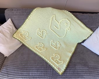 May include: A pale yellow crochet blanket featuring a repeating pattern of raised bobble stitch ducks. The blanket has a textured bobble stitch border in the same pale yellow color and a small rectangular tag attached to the bottom corner.
