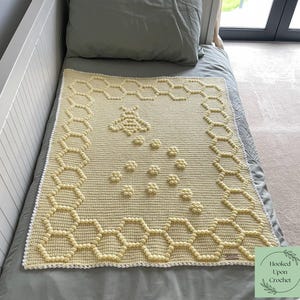 May include: A handmade, light yellow crochet blanket with a bee and honeycomb design. The blanket features a repeating honeycomb pattern around the edges and a bee and flower motif in the center. The blanket is displayed on a bed.