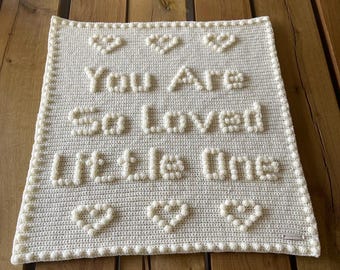 Puede incluir: Una manta rectangular de ganchillo color crema con textura de punto burbuja. El texto "You Are So loved little One" está centrado en la manta, rodeado por seis formas de corazón, tres en la parte superior y tres en la inferior, todas en punto burbuja color crema.