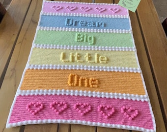 Puede incluir: Una manta de ganchillo rectangular con rayas horizontales en rosa pastel, azul, verde, amarillo y naranja. La manta muestra el texto Dream Big Little One en puntos de bodoque en relieve, con filas de corazones de bodoque arriba y abajo.