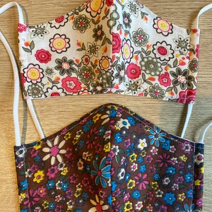 May include: Two face coverings with floral patterns and white elastic ear loops. One features a white base with pink, yellow, and green flowers. The other has a brown base with blue, pink, and yellow flowers.