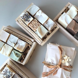 May include: Several wooden boxes filled with various handmade soaps. The soaps are different colors and textures, some with dried lavender. One wrapped package is tied with a ribbon and dried flowers. The soaps are arranged on a light-colored surface.