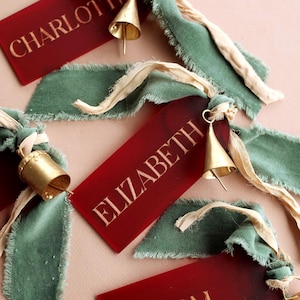 May include: Rectangular red acrylic tags with gold engraved names including CHARLOTTE and ELIZABETH. Each tag is attached to a frayed sage green velvet ribbon and a cream-colored silk ribbon, accented with a small metallic gold bell.