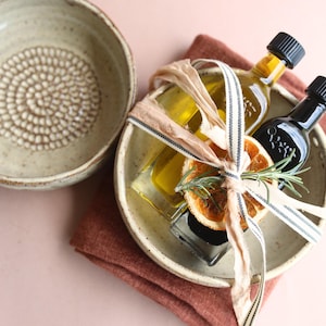May include: A ceramic bowl with a textured interior sits next to a bowl containing two glass bottles of oil and vinegar, tied with ribbon and decorated with a dried orange slice and rosemary sprigs. The items rest on a rust-colored cloth.