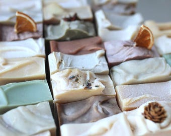 May include: An assortment of handmade soaps in various colors and textures. Some soaps are topped with dried orange slices, lavender, or a small pine cone. The soaps are arranged in a grid pattern, showcasing their unique designs and natural ingredients.
