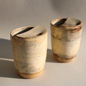 May include: Two ceramic mugs with a speckled, off-white glaze and a brown rim. The mugs have a unique, asymmetrical lip.