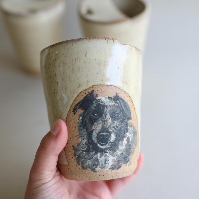 May include: A ceramic mug with a light brown glaze and a black and white dog portrait painted on the side. The dog has a black nose and white fur around its eyes.