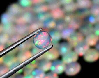May include: A single iridescent opal gemstone held by tweezers, surrounded by a scattering of similar opals.
