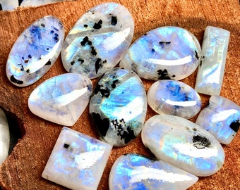 May include: A collection of iridescent rainbow moonstone cabochons with black inclusions. The stones are various shapes and sizes and are arranged on a piece of wood.