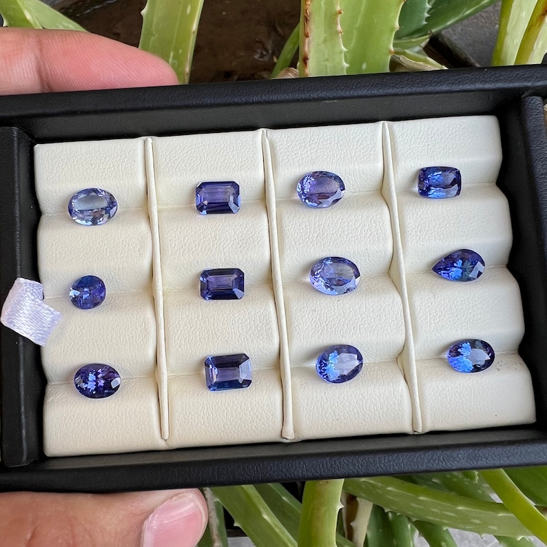 May include: A collection of twelve loose gemstones in various shapes and sizes, including oval, pear, and emerald cuts, displayed in a white velvet-lined box with black trim.