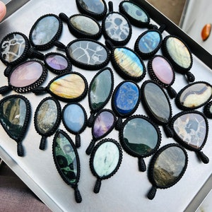 Peut inclure: Une collection de pendentifs en pierres pr&eacute;cieuses, chacun envelopp&eacute; d'un cordon en macram&eacute; noir. Les pierres sont un m&eacute;lange de couleurs et de motifs, notamment le bleu, le vert, le violet, le rose et le brun.