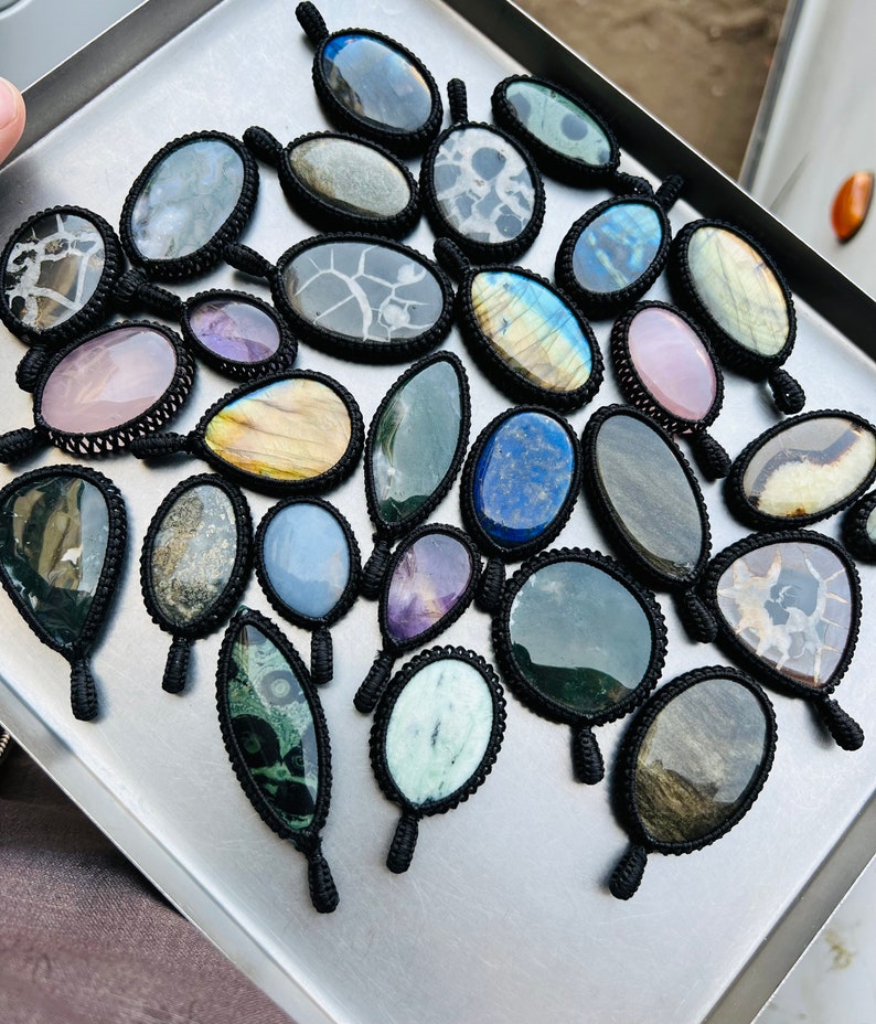 Peut inclure: Une collection de pendentifs en pierres pr&eacute;cieuses, chacun envelopp&eacute; d'un cordon en macram&eacute; noir. Les pierres sont un m&eacute;lange de couleurs et de motifs, notamment le bleu, le vert, le violet, le rose et le brun.
