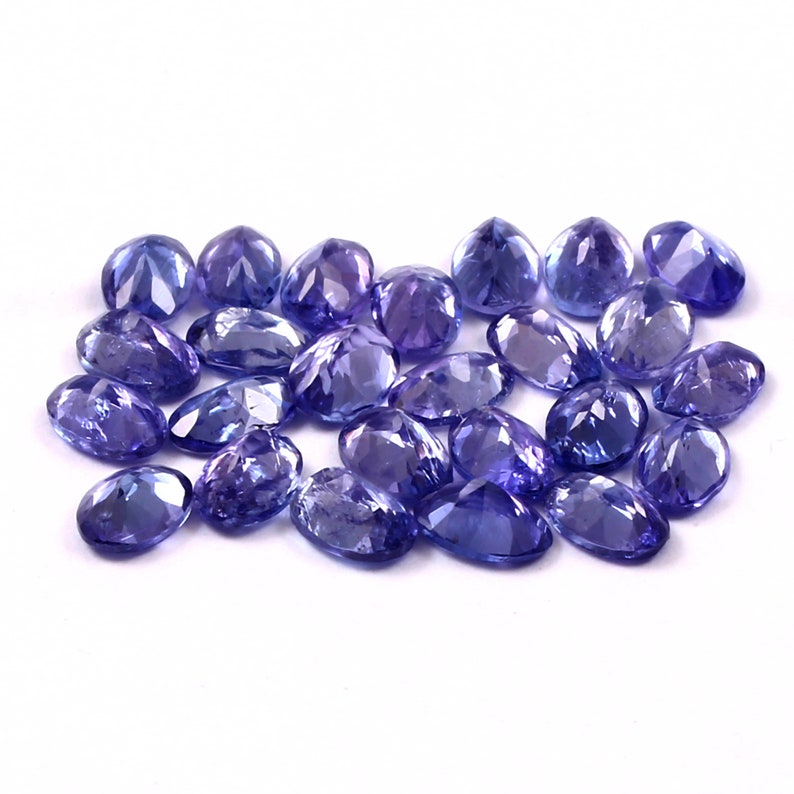 Puede incluir: Una colecci&oacute;n de piedras preciosas ovaladas facetadas en tonos de p&uacute;rpura y azul. Las piedras preciosas est&aacute;n dispuestas en un mont&oacute;n suelto sobre un fondo blanco.