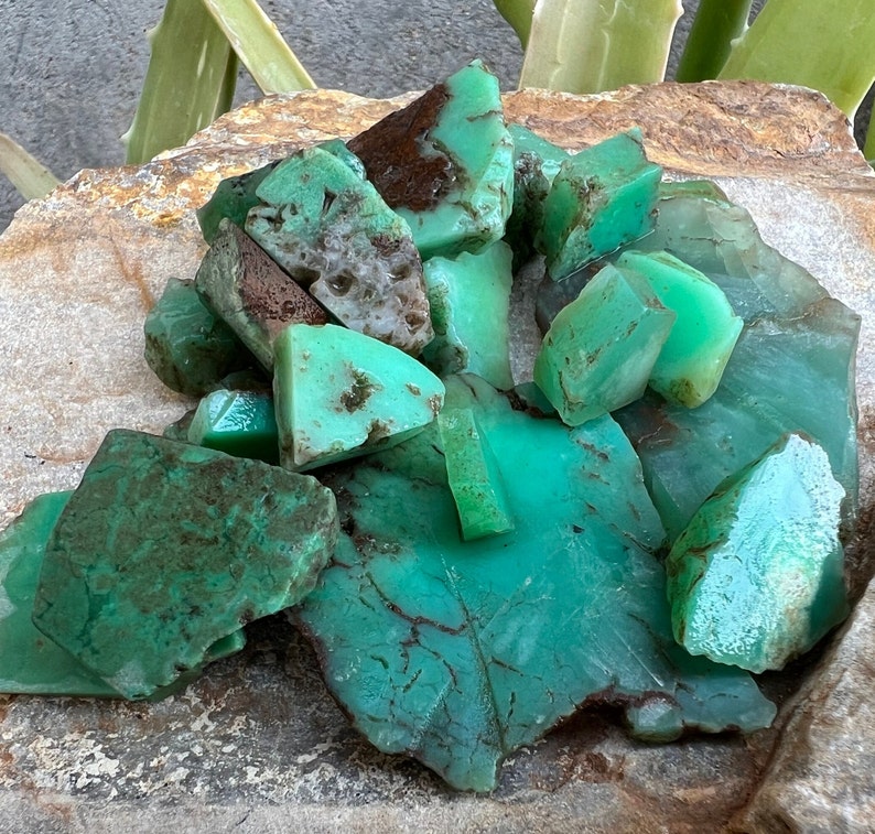 May include: A collection of green and brown chrysoprase gemstones, some with a rough texture, others with a smooth surface. The stones are arranged on a brown, textured surface.