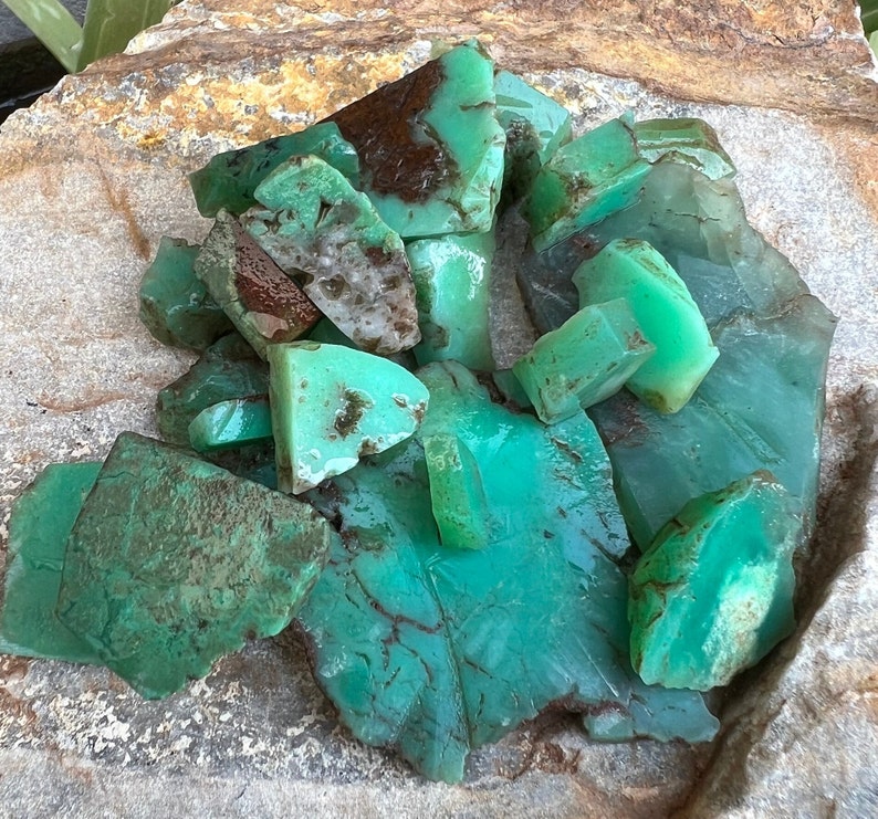 May include: A collection of green and brown chrysoprase gemstones, with a variety of shapes and sizes, displayed on a rough, textured surface.