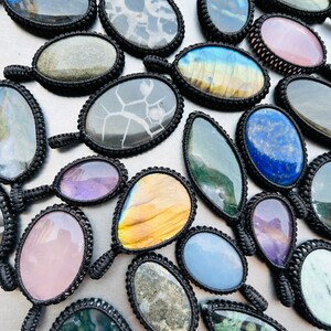 Peut inclure: Une collection de pendentifs en pierres pr&eacute;cieuses de forme ovale et en forme de larme avec des enroulements de cordon noir. Les pierres sont de diff&eacute;rentes couleurs, notamment le vert, le bleu, le violet, le rose et le brun.