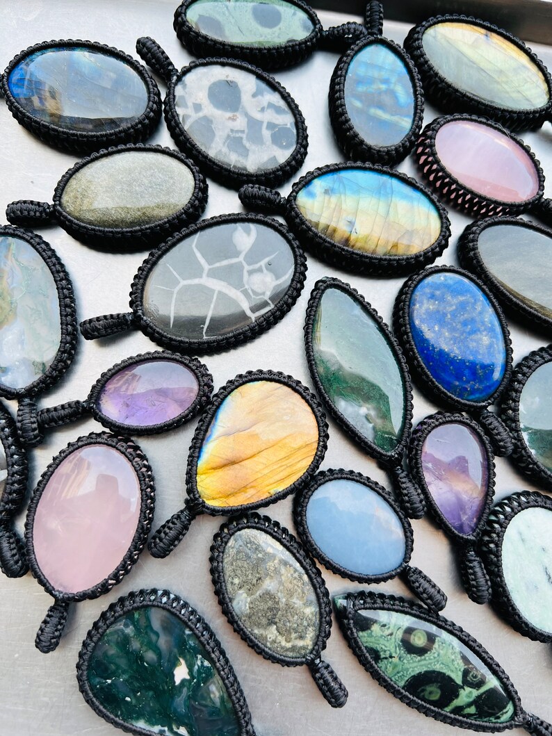Peut inclure: Une collection de pendentifs en pierres pr&eacute;cieuses de forme ovale et en forme de larme avec des enroulements de cordon noir. Les pierres sont de diff&eacute;rentes couleurs, notamment le vert, le bleu, le violet, le rose et le brun.