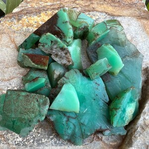 May include: A collection of green and brown chrysoprase gemstones, some with a rough texture, in a pile on a light brown surface.