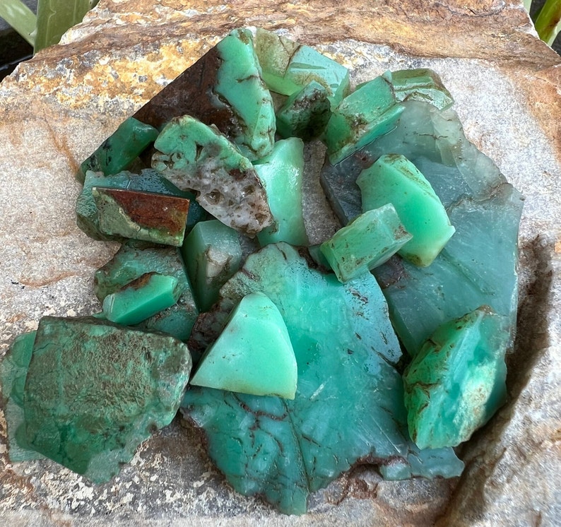 May include: A collection of green and brown chrysoprase gemstones, some with a rough texture, in a pile on a light brown surface.
