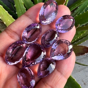 May include: A collection of eight amethyst gemstones, cut into pear and oval shapes, with a purple hue. The gemstones are displayed on a hand.