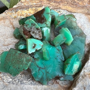 May include: A collection of green and brown chrysoprase gemstones, some with a rough texture, on a light brown rock surface.