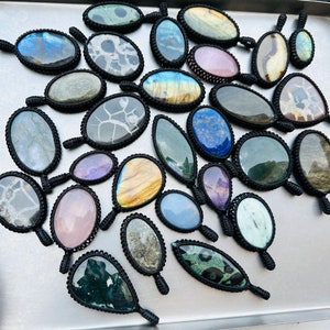 Peut inclure: Assortiment de pendentifs en pierres pr&eacute;cieuses color&eacute;es avec des enroulements de cordon noir. Chaque pendentif pr&eacute;sente une pierre unique, y compris le bleu, le vert, le violet et le brun.