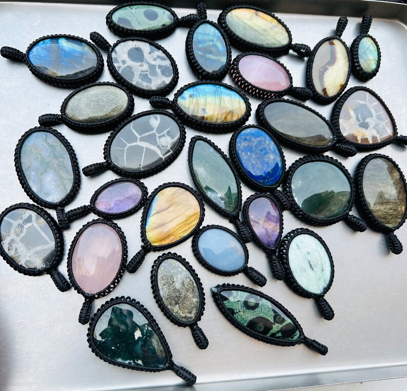 Peut inclure: Assortiment de pendentifs en pierres pr&eacute;cieuses color&eacute;es avec des enroulements de cordon noir. Chaque pendentif pr&eacute;sente une pierre unique, y compris le bleu, le vert, le violet et le brun.