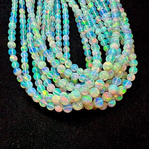 Puede incluir: Hilos de cuentas de ópalo iridiscentes. Las cuentas son redondas y muestran una gama de colores, incluyendo azul, verde y amarillo. Las cuentas están ensartadas, creando un efecto de capas. El fondo es negro.