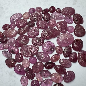 May include: A selection of carved gemstones in shades of pink and purple, with floral and leaf patterns. The gemstones are in various shapes, including oval and round, and have polished surfaces. The collection is a mix of sizes and tones.
