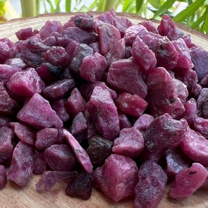 Può includere: Un mucchio di pietre preziose di rubino grezze di varie dimensioni e forme. Le pietre non tagliate mostrano un colore rosso-viola intenso, con alcune aree più scure. Le pietre preziose sono sparse su una superficie di legno, mostrando il loro stato naturale non lucidato.