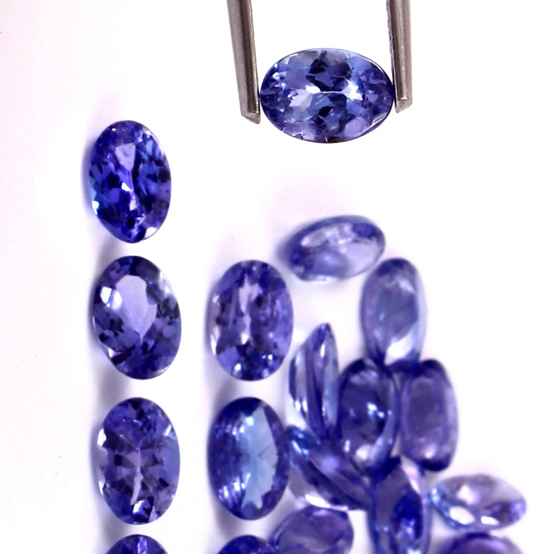 Puede incluir: Una colecci&oacute;n de piedras preciosas azules de forma ovalada y facetadas. Las piedras preciosas est&aacute;n sueltas y dispersas sobre una superficie blanca. Una piedra preciosa est&aacute; siendo sostenida por unas pinzas.