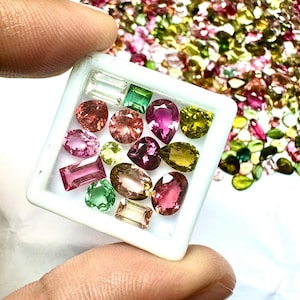 May include: A collection of colorful gemstones in a white tray, held by a hand. The gems are various shapes and sizes, including oval, rectangular, and pear-shaped. Colors include pink, green, yellow, and purple. A scattering of loose gems is in the background.